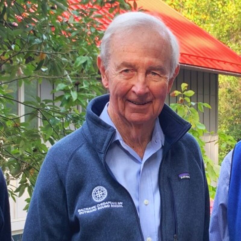The image shows an older man with white hair, wearing a blue jacket over a light blue shirt. He is smiling and appears to be outdoors, with a building and greenery in the background. The jacket has a logo on it. The man seems to be in a relaxed setting.